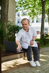 Fototapeta premium A man with inclusivity sits on a bench in a park, using a laptop computer.