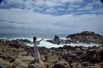 portrait of female model  standing wearing white goddess dress, dramatic natural landscape background of rocky ocean shoreline with stone clifftops. castle rock, Busselton, Western Australia