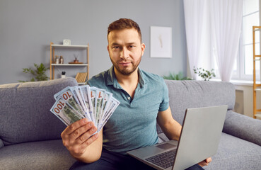 Concentrated rich man seated on a couch at home, holding stack cash and using a laptop, celebrating...