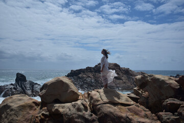 portrait of female model  standing wearing white goddess dress, dramatic natural landscape background of rocky ocean shoreline with stone clifftops. castle rock, Busselton, Western Australia