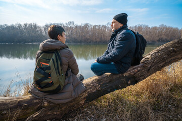 Two men sitting on a log by the river, father and son, early spring landscape, hiking and outdoor...
