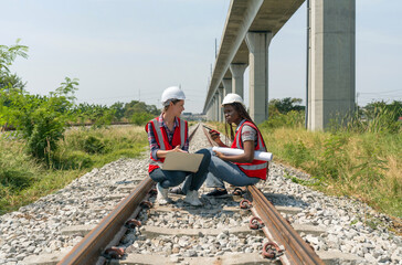 Two engineer with safety helmet and high-visibility vest reviewing work to be done on a railway track project while holding a set of plans or blueprint, walkie-talkie and laptop computer.