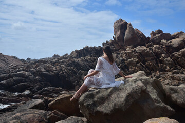 portrait of female model sitting wearing white goddess dress, dramatic natural landscape background of rocky ocean shoreline with stone clifftops. castle rock, Busselton, Western Australia