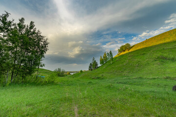 A rolling, green hillside with a small path leading into the distance, framed by lush trees and a sunlit sky.