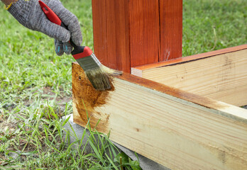 A hand wearing a glove applies a stain to the bottom of a wooden structure with a paintbrush. The...