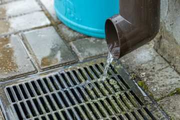 Water flows from a brown gutter into a metal drain grate, creating a small stream. The drain is surrounded by grey pavement stones and green moss.