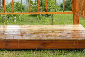 A wooden deck patio in a backyard, with rain falling and reflecting off the wet surface. The deck...