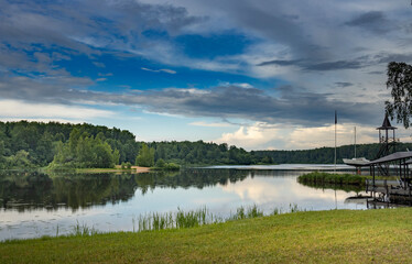 A calm lake reflecting a dramatic sky, with lush green trees lining the shore and a small wooden cabin nestled on the waters edge..