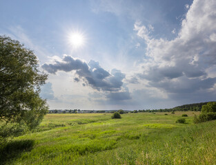 Obraz premium A wide shot of a verdant field under a partly cloudy spring sky. The sun shines brightly through the clouds, casting long shadows over the landscape.