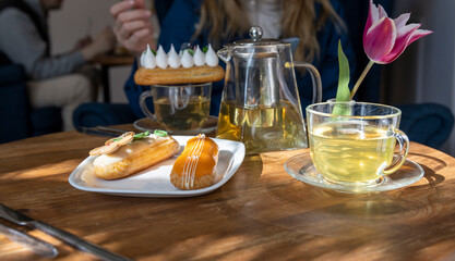 Enjoy a serene afternoon tea setting with a variety of pastries, including eclairs, served on a wooden table bathed in sunlight. The ambiance is complemented by a vibrant tulip