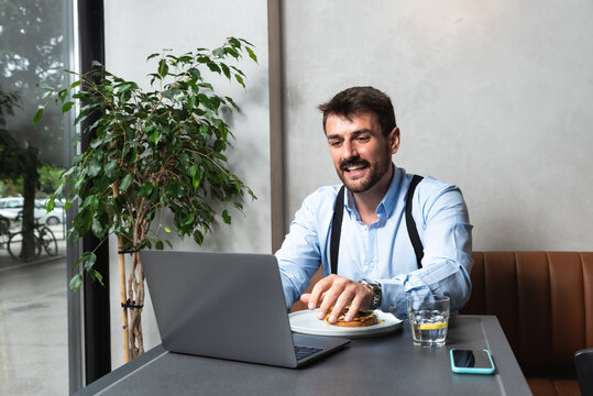Young hungry business man at lunch break sitting at cafeteria eating hamburger and using laptop for online work. Burger lover business person eat and working from restaurant.