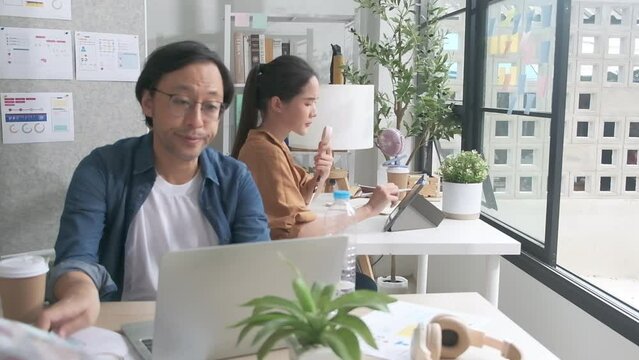 Two Asian workers are unhappy working with hot temperatures at casual office in summer season, uncomfortable with no air conditioner, using fans and drinking water, sitting at desks, and sweating.