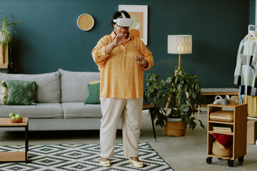 Senior African American woman immersing herself in gaming process thanks to VR goggles and standing in middle of living room