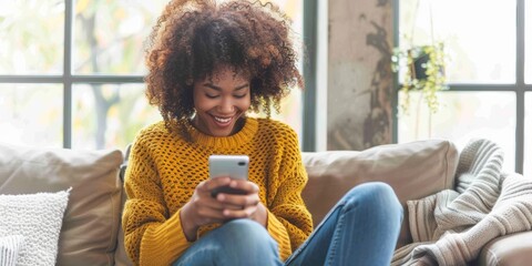 At home, a joyful mixed-race woman drinks coffee and types on her phone. One happy Hispanic woman with a curly afro uses her phone for social media on the couch at home.