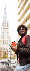 Smiling indian man in sunglasses holding coffee and smartphone on street, vertical banner 