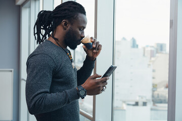 An African Man with dreadlocks is drinking coffee while looking at his cell phone reading messages