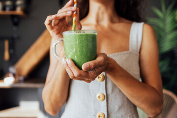 A transparent cup with a green smoothie in the hands of a young woman. A girl in a linen sundress...