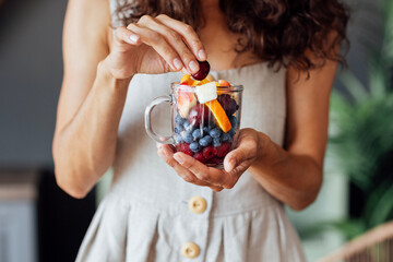 Close-up of a glass mug with fruits and berries in a female well-groomed hands. A young woman in a sundress is eating cherries at home.