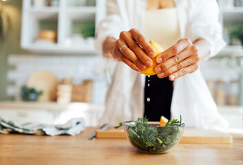 A female well-groomed hands with a neat manicure, squeezing lemon juice into a glass bowl with salad. Healthy food. Fresh green salad with arugula, onion and parsley.