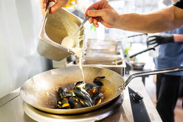 Person preparing a seafood dish, pouring creamy sauce into a pan of mussels on a stove, in a kitchen setting