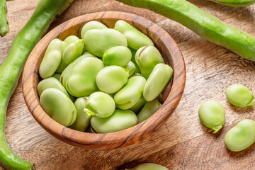 Broad beans in wooden bowl and broad bean pods near it on wooden table. Top view.