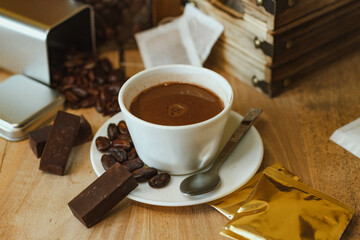 Cocoa bean tea packet in a white glass on a wooden table with chocolate chunks next to it surrounded by seeds.