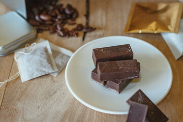 Cocoa bean tea packet in a white glass on a wooden table with chocolate chunks next to it surrounded by seeds.