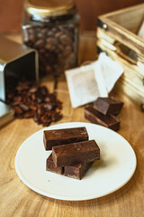 Cocoa bean tea packet in a white glass on a wooden table with chocolate chunks next to it surrounded by seeds.