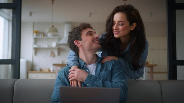 Caucasian couple family at home man guy freelancer male husband boyfriend using laptop on couch sofa girl female woman girlfriend wife going from kitchen hugging back talking discuss computer shopping