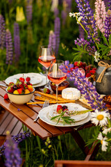 Beautiful elegant outdoor arrangement in meadow with purple blue lupins. Perfect date, private wedding or romantic marriage proposal. Summer, sunset, golden hour. Appetisers, rose wine, strawberry