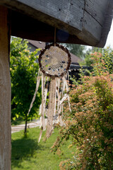 A boho style dreamcatcher hangs from a wooden cabin with a flowering garden as a backdrop.