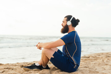 A man with a beard relaxes on a beach, listening to music with headphones, enjoying the sunny outdoors