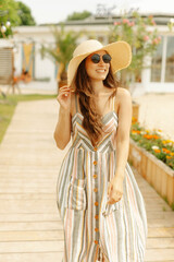 A woman is smiling in a sundress and straw hat as she strolls on a flowerlined wooden pathway on a sunny day