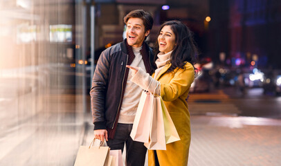 A couple standing in front of a store, holding shopping bags filled with purchases. They look happy...