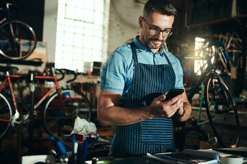 Bicycle, garage and phone for man, reading and small business of mechanic, apron and equipment. Proud, owner and person with service, maintenance and tools for bike, technician or working with mobile
