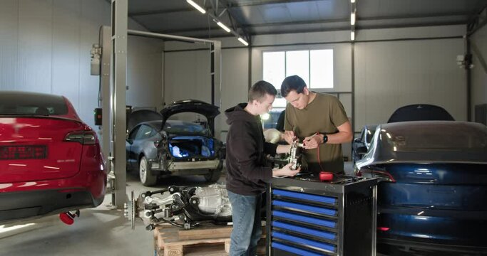 EV repair concept. Auto Service. Two male mechanics working together on an electric car engine repair in a modern automobile workshop. Specialized tools and electric vehicles visible in the background