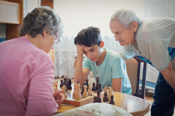 Selective focus thoughtful young boy plays chess with his grandparents, creating a moment of bonding and strategy set in a cozy indoor environment. International Chess Day.