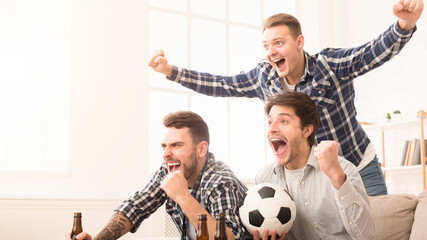 Three enthusiastic men celebrate with cheers and raised hands, holding a soccer ball and beer...