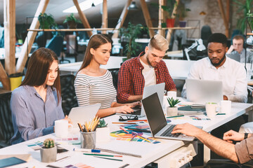 A group of young professionals work together in a modern, open-office environment with natural light, laptops, and design materials.