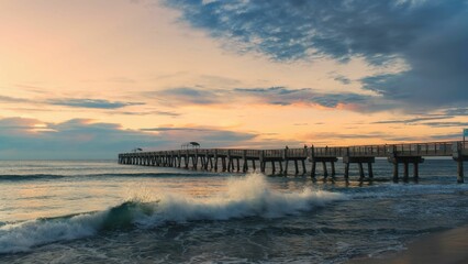 Obraz premium Scenic view of a pier extending into the ocean at sunset with waves crashing on the shore.