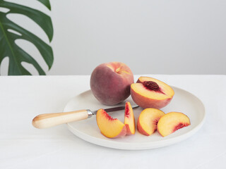 White plate with knife on the table with ripe peaches cut into slices and halves. Green plant leaf in the background.