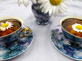 Porcelain blue cups with saucers, decorated with gilding, filled with chamomile herbal tea, vase with fresh chamomiles stand in a row on a light blue tablecloth