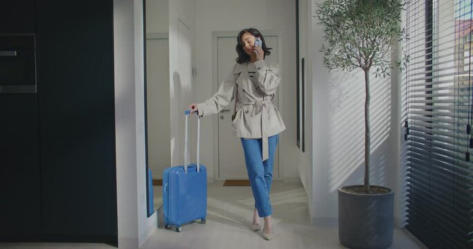Cute Caucasian female talking on smartphone. Standing next to house entrance. Leaning on expandable handle of suitcase. Talking on smartphone before leaving. Autumn outfit. Communication concept.