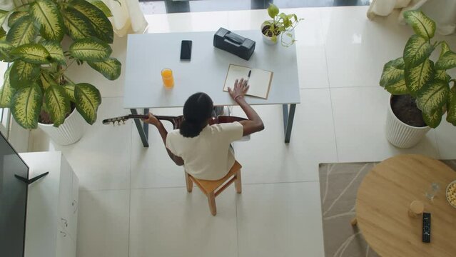 Directly above view of black woman playing guitar when writing song using notebook sitting at table at home