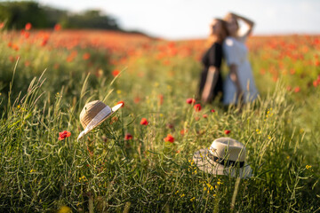 Zwei Hüte im Vordergrund mit zwei Frauen im Hintergrund in einer Wiese mit Mohnblumen