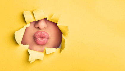 A close-up view of a woman lips subtly peeking out of a hole in a bright yellow paper. The lips are...