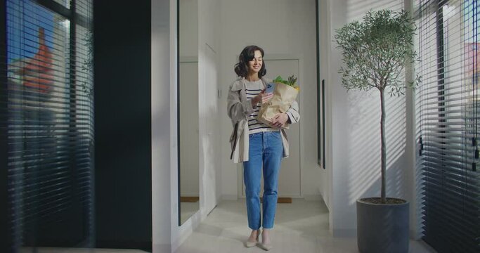 Caucasian brunette standing in middle of lobby. Holding bag of groceries with one hand. Wearing jeans, stylish jacket and high heels. Using mobile device. Typing message to husband or boyfriend.