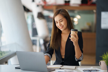 Young professional woman working on laptop at modern office while holding a coffee cup....