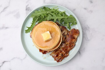 Tasty pancakes with butter, fried bacon and fresh arugula on white marble table, top view