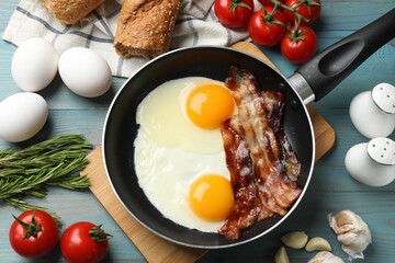 Tasty bacon and eggs in frying pan among products on light blue table, flat lay